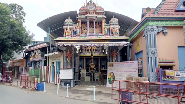 A Hindu temple with intricate carvings and colorful statues is set in an urban environment. The entrance features traditional architectural elements with brightly painted figures of deities. A canopy extends over the gate, and informative signs are displayed near the entrance. The surroundings include red metal barricades and a large tree to the side.