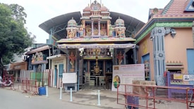 A Hindu temple with intricate carvings and colorful statues is set in an urban environment. The entrance features traditional architectural elements with brightly painted figures of deities. A canopy extends over the gate, and informative signs are displayed near the entrance. The surroundings include red metal barricades and a large tree to the side.