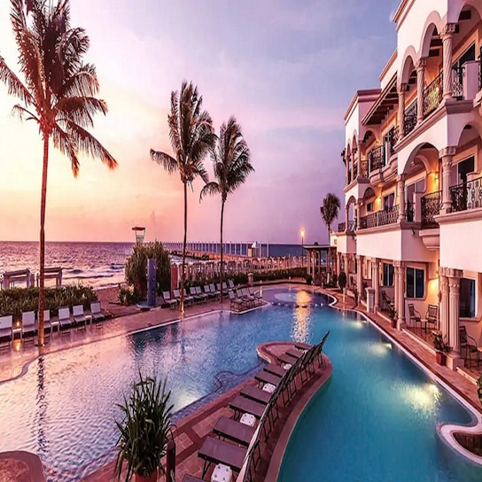 A luxurious resort setting with a large swimming pool surrounded by lounge chairs, palm trees, and a grand building with balconies. The sun is setting over the ocean in the background, casting a warm, pinkish hue over the entire scene.