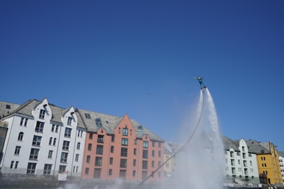 A person is flyboarding over water, elevated by a powerful jet stream. In the background, colorful buildings with gabled roofs line the waterfront. The sky is clear and blue.