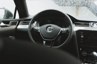 Interior view of a car focusing on the steering wheel with a Volkswagen logo. The dashboard and part of the center console are visible. The windows are covered in raindrops, indicating rainy weather outside.