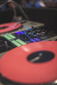 A close-up shot of a DJ mixer and turntables with vibrant red vinyl records. The mixer features various knobs, buttons, and sliders, including illuminated green and blue buttons, creating a dynamic and energetic atmosphere.