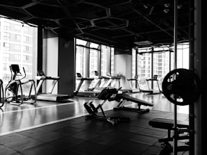 A modern gym with an array of exercise equipment such as treadmills, elliptical machines, and weight benches. Large windows along the wall allow natural light to illuminate the space. The floor is equipped with rubber mats for safety and comfort.