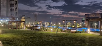 A nighttime view of a landscaped area with well-maintained lawns, illuminated pathways, and small trees. There are pergolas and structures with soft lighting surrounding a blue-lit pool. High-rise buildings are visible in the background under a cloudy sky.