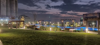 A nighttime view of a landscaped area with well-maintained lawns, illuminated pathways, and small trees. There are pergolas and structures with soft lighting surrounding a blue-lit pool. High-rise buildings are visible in the background under a cloudy sky.
