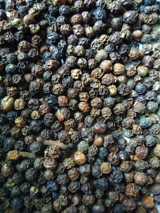 A close-up view of a large quantity of dried black peppercorns with some cloves scattered among them. The peppercorns vary in shades of dark brown, from almost black to lighter brown hues.