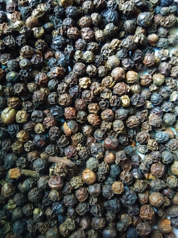 A close-up view of a large quantity of dried black peppercorns with some cloves scattered among them. The peppercorns vary in shades of dark brown, from almost black to lighter brown hues.