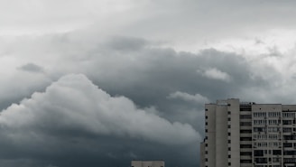 A large, dense cloud formation looms over a tall, modern building. The sky appears overcast with varying shades of gray and hints of darker, stormy clouds. The building features numerous windows, suggesting a residential or office structure.