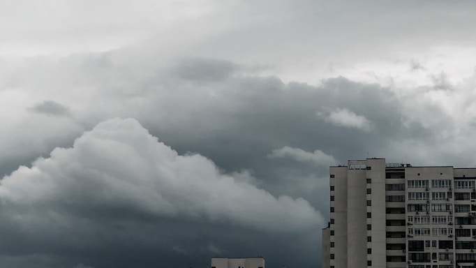 A large, dense cloud formation looms over a tall, modern building. The sky appears overcast with varying shades of gray and hints of darker, stormy clouds. The building features numerous windows, suggesting a residential or office structure.