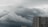 A large, dense cloud formation looms over a tall, modern building. The sky appears overcast with varying shades of gray and hints of darker, stormy clouds. The building features numerous windows, suggesting a residential or office structure.