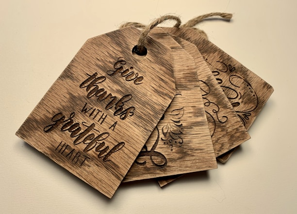 A set of four rustic wooden tags with string, featuring engraved calligraphy and decorative designs. The front tag displays the phrase 'Give thanks with a grateful heart'. The wood has a natural, textured finish.