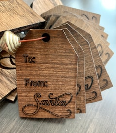 A stack of wooden gift tags with dark engraving, featuring spaces labeled 'To' and 'From Santa' with a decorative script for 'Santa'. The tags are tied together with brown string and a gold metal spiral decoration.