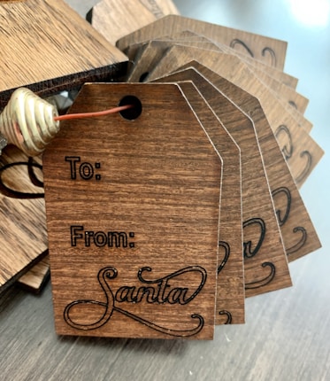 A stack of wooden gift tags with dark engraving, featuring spaces labeled 'To' and 'From Santa' with a decorative script for 'Santa'. The tags are tied together with brown string and a gold metal spiral decoration.