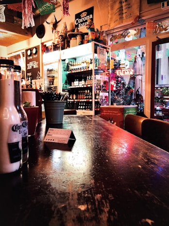 A rustic bar setting featuring a wooden counter with a slightly worn finish. Various bottles are visible on a shelf against the wall, and a prominent Corona Extra sign is displayed above. Decor includes festive paper cut-outs suspended from the ceiling and an arrangement of items like a plant in a pot and bottles on the counter.