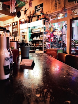 A rustic bar setting featuring a wooden counter with a slightly worn finish. Various bottles are visible on a shelf against the wall, and a prominent Corona Extra sign is displayed above. Decor includes festive paper cut-outs suspended from the ceiling and an arrangement of items like a plant in a pot and bottles on the counter.