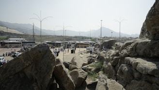 A group of buses is parked on a flat area surrounded by rocky terrain and mountains in the background. People in light-colored clothing are gathered in small clusters around the area. Large rocks dominate the foreground, while tall lamp posts are scattered throughout the scene.