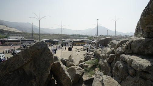 A group of buses is parked on a flat area surrounded by rocky terrain and mountains in the background. People in light-colored clothing are gathered in small clusters around the area. Large rocks dominate the foreground, while tall lamp posts are scattered throughout the scene.
