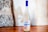 A bottle with a frosted design stands on a wooden surface. It has a blue cap and a label featuring artwork and the word 'amarguinha'. The background consists of a textured, rustic wall with a mix of stone and plaster.