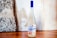 A bottle with a frosted design stands on a wooden surface. It has a blue cap and a label featuring artwork and the word 'amarguinha'. The background consists of a textured, rustic wall with a mix of stone and plaster.