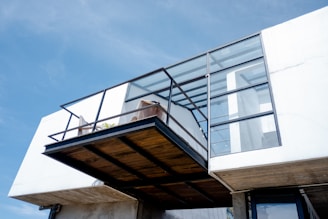 A modern architectural structure featuring a cantilevered balcony with metal and glass elements. The balcony area contains a few potted plants and a minimalistic design. The building has a clean, white façade with large glass windows and panels, allowing ample natural light to enter.