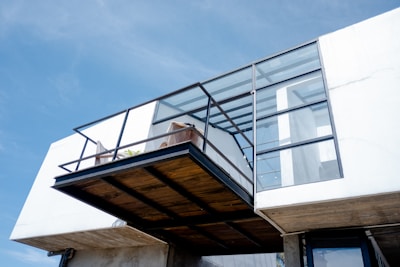 A modern architectural structure featuring a cantilevered balcony with metal and glass elements. The balcony area contains a few potted plants and a minimalistic design. The building has a clean, white façade with large glass windows and panels, allowing ample natural light to enter.
