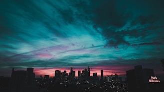 A city skyline with a silhouette of tall buildings is set against a dramatic sunset. The sky features striking hues of teal, pink, and purple, with dense cloud formations adding texture to the scene. A sign on one of the buildings reads 'Park Wilshire'.