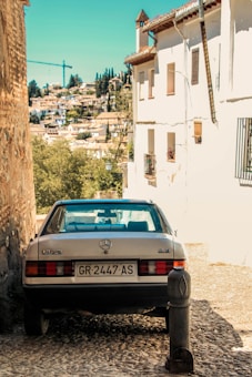 A vintage Mercedes-Benz car is parked along a narrow cobblestone street. The area is lined with whitewashed buildings featuring wrought-iron balconies. In the background, a cityscape with a mix of historic and modern structures surrounded by lush greenery is visible. A crane can be seen in the distance, indicating ongoing construction.