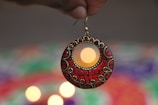 A hand holds an intricately designed earring with red and black patterns and gold accents. In the background, blurred lights and vibrant colors create a visually appealing contrast.