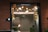 A single lit diya is placed on the left side of the foreground, casting a warm glow. The background is softly blurred, showcasing decorative lights and garlands hanging across an entranceway. The setting appears festive and tranquil.