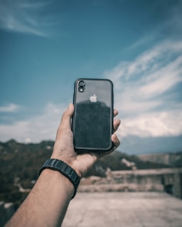 A hand holding a smartphone with a clear case that displays an apple logo, suggesting it is an Apple device. The background features a clear blue sky with some clouds and a distant blurry landscape.
