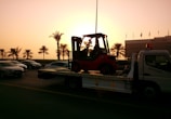 A sunset or sunrise scene with a tow truck carrying a forklift on a highway. The silhouette of palm trees is visible in the background, along with several cars on the road. A building is visible on the right side with a row of flags on top.