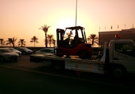 A sunset or sunrise scene with a tow truck carrying a forklift on a highway. The silhouette of palm trees is visible in the background, along with several cars on the road. A building is visible on the right side with a row of flags on top.