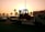 A sunset or sunrise scene with a tow truck carrying a forklift on a highway. The silhouette of palm trees is visible in the background, along with several cars on the road. A building is visible on the right side with a row of flags on top.