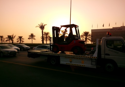 A sunset or sunrise scene with a tow truck carrying a forklift on a highway. The silhouette of palm trees is visible in the background, along with several cars on the road. A building is visible on the right side with a row of flags on top.