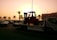 A sunset or sunrise scene with a tow truck carrying a forklift on a highway. The silhouette of palm trees is visible in the background, along with several cars on the road. A building is visible on the right side with a row of flags on top.
