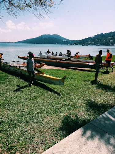 A group of people gather on a grassy shore beside several colorful canoes. The canoes are positioned near the water, and in the background, a blue sea stretches out to a mountainous landscape under a clear sky. Some individuals are dressed in bright clothing, indicating a possible water sports activity.