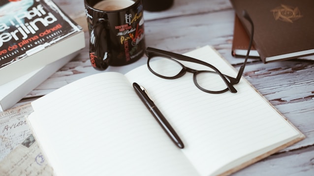 An open notebook sits on a wooden table, flanked by a pair of eyeglasses and a pen resting on its pages. Nearby, a coffee mug with a colorful design holds a beverage. In the background, there are books and a journal adding to the cozy, intellectual atmosphere.