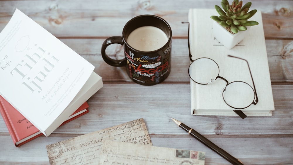 Taza de cerámica negra y multicolor con líquido blanco junto a papeles, libros y anteojos