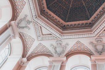An ornate architectural ceiling with intricate designs, featuring octagonal patterns and elaborate moldings. The ceiling includes decorative plasterwork and cornices, with arched windows visible along the edges.