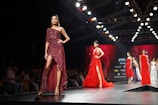 A fashion runway featuring several models walking in glamorous, sparkling dresses. The scene is illuminated by overhead lights with a focus on a model wearing a sequined, off-shoulder dress in deep red. The background is a large digital screen with red patterns.