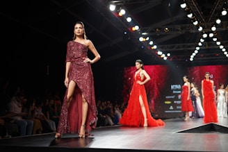A fashion runway featuring several models walking in glamorous, sparkling dresses. The scene is illuminated by overhead lights with a focus on a model wearing a sequined, off-shoulder dress in deep red. The background is a large digital screen with red patterns.