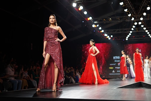 A fashion runway featuring several models walking in glamorous, sparkling dresses. The scene is illuminated by overhead lights with a focus on a model wearing a sequined, off-shoulder dress in deep red. The background is a large digital screen with red patterns.