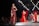 A fashion runway featuring several models walking in glamorous, sparkling dresses. The scene is illuminated by overhead lights with a focus on a model wearing a sequined, off-shoulder dress in deep red. The background is a large digital screen with red patterns.