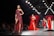 A fashion runway featuring several models walking in glamorous, sparkling dresses. The scene is illuminated by overhead lights with a focus on a model wearing a sequined, off-shoulder dress in deep red. The background is a large digital screen with red patterns.