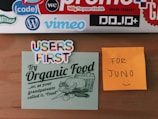 A wooden surface displays several colorful stickers and notes. At the top, there are various tech and brand stickers like 'bitly', 'vimeo', and 'code'. Below them, two notes are stuck to the surface: one is a light green card with the phrase 'Try Organic Food' accompanied by a vintage illustration of a basket of vegetables; another is a bright orange sticky note with the handwritten message 'FOR JUNO :)'. Additionally, there's a sticker with 'USERS FIRST' written in multicolored letters.
