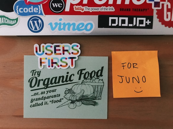 A wooden surface displays several colorful stickers and notes. At the top, there are various tech and brand stickers like 'bitly', 'vimeo', and 'code'. Below them, two notes are stuck to the surface: one is a light green card with the phrase 'Try Organic Food' accompanied by a vintage illustration of a basket of vegetables; another is a bright orange sticky note with the handwritten message 'FOR JUNO :)'. Additionally, there's a sticker with 'USERS FIRST' written in multicolored letters.