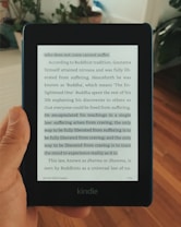 A hand holding a Kindle displaying a page of text. The text appears to be from a book discussing Buddhist teachings, particularly focusing on the concept that suffering arises from craving.