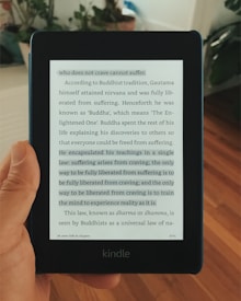 A hand holding a Kindle displaying a page of text. The text appears to be from a book discussing Buddhist teachings, particularly focusing on the concept that suffering arises from craving.