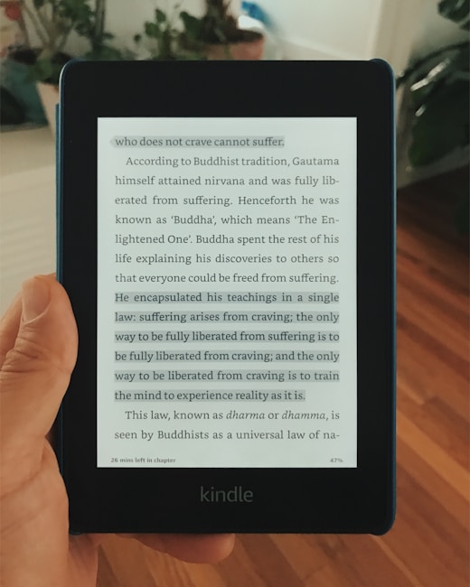 A hand holding a Kindle displaying a page of text. The text appears to be from a book discussing Buddhist teachings, particularly focusing on the concept that suffering arises from craving.