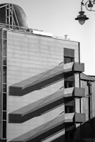 A monochromatic, industrial-style building facade with external metal staircases casting long, geometric shadows on the flat, tiled surface. A metallic dome and a vintage streetlamp add contrast to the modern architectural lines.